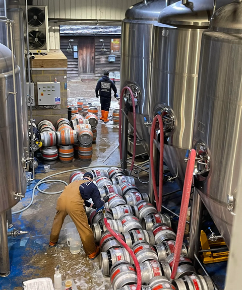 Brewery interior with large stainless steel tanks and workers handling kegs.