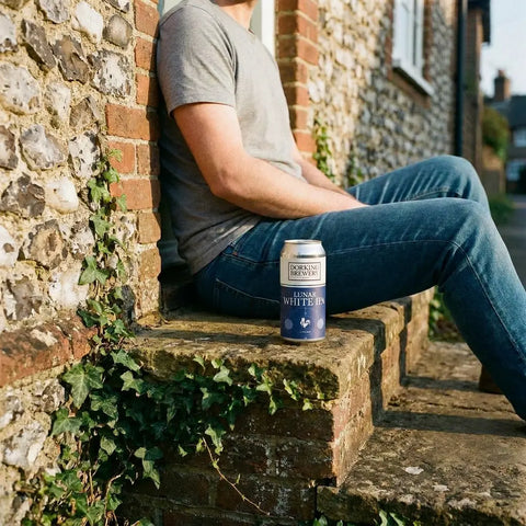 A can of Dorking Brewery Lunar White IPA on stone steps beside a person resting against a flint wall.