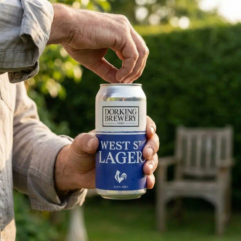 Person opening a can of West St Lager beer with a blurred outdoor background